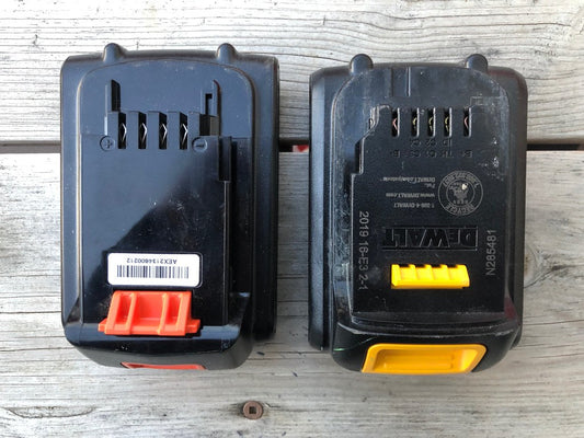 Two power tool battery packs side by side on a wooden surface, one with an orange release button and one DeWalt battery with a yellow tab